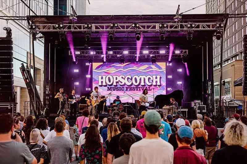 A band performing on a stage in front of an audience in Raleigh with the sign "Hopscotch Music Fest" behind them.